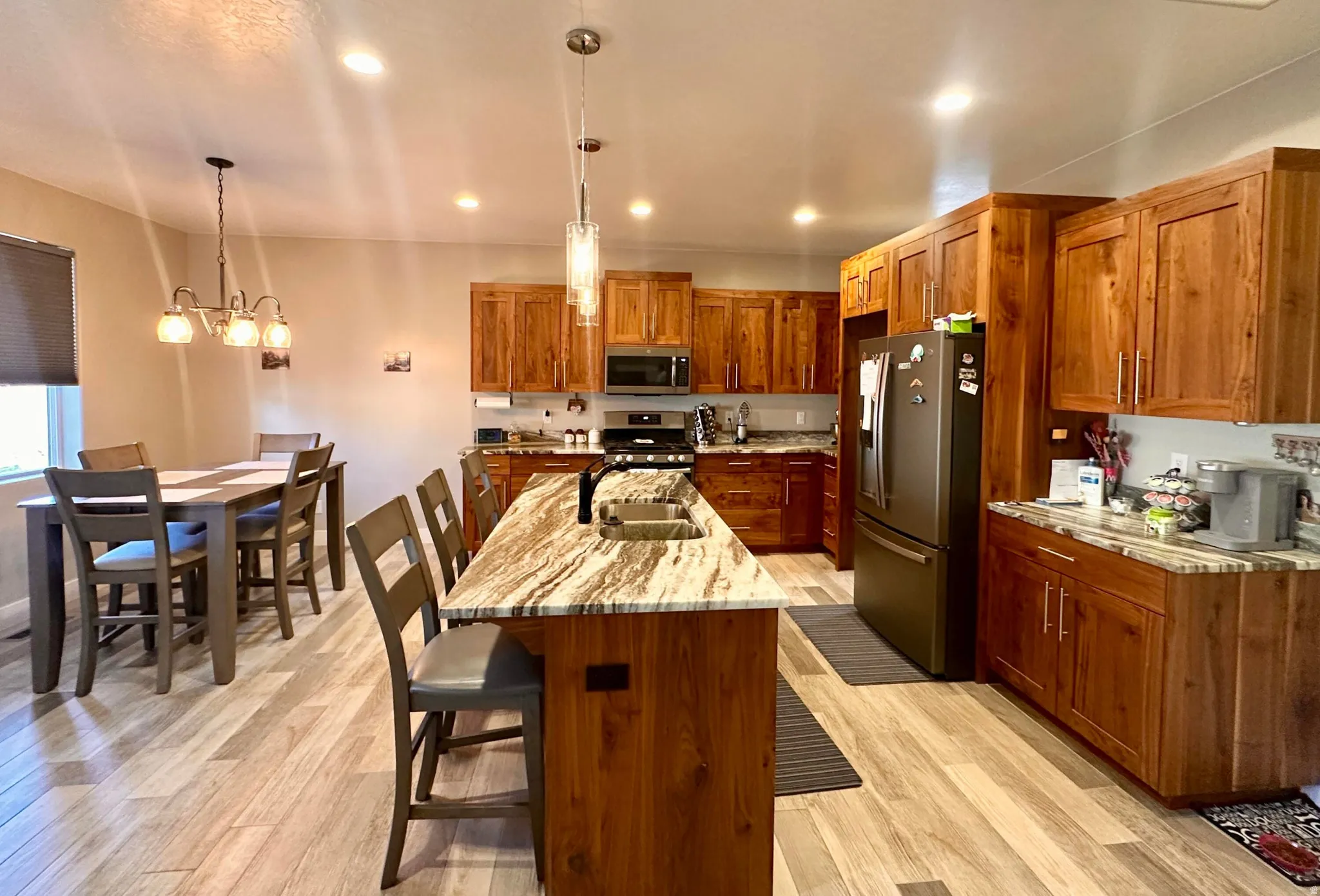 Kitchen featuring brown cabinets, light stone counters, stainless steel appliances, pendant lighting, and a kitchen island with sink