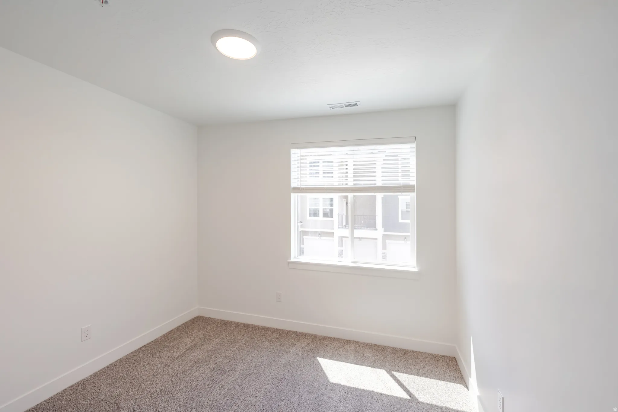 Bright interior room featuring a single window with horizontal blinds, neutral-toned carpet flooring, white painted walls, and a ceiling-mounted flush light fixture