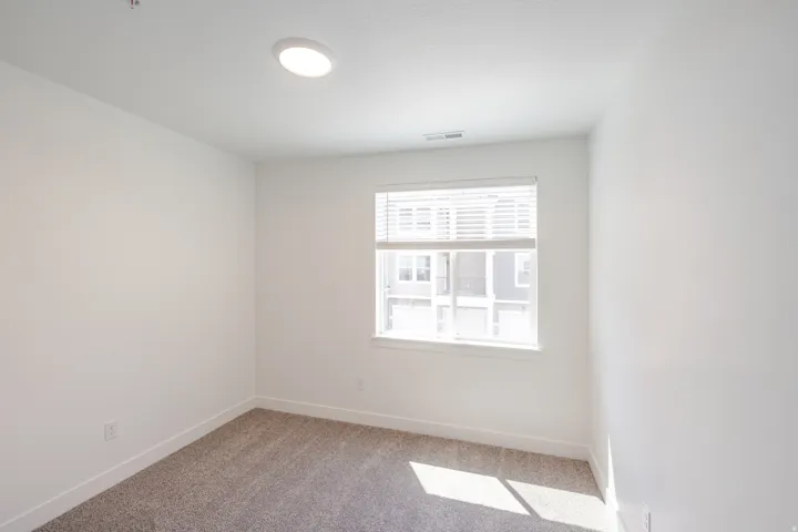 Bright interior room featuring a single window with horizontal blinds, neutral-toned carpet flooring, white painted walls, and a ceiling-mounted flush light fixture