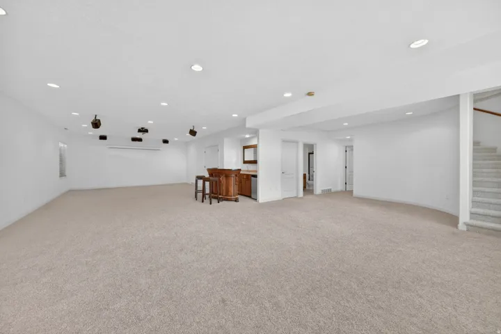Spacious carpeted room featuring recessed lighting, an integrated home theater system with screen and speakers, a built-in wet bar with wooden cabinetry, and a carpeted staircase with a wood handrail