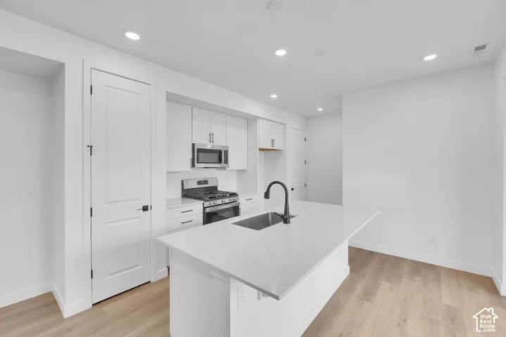 Kitchen with white cabinets, stainless steel appliances, an island with sink, light wood finished floors, and recessed lighting