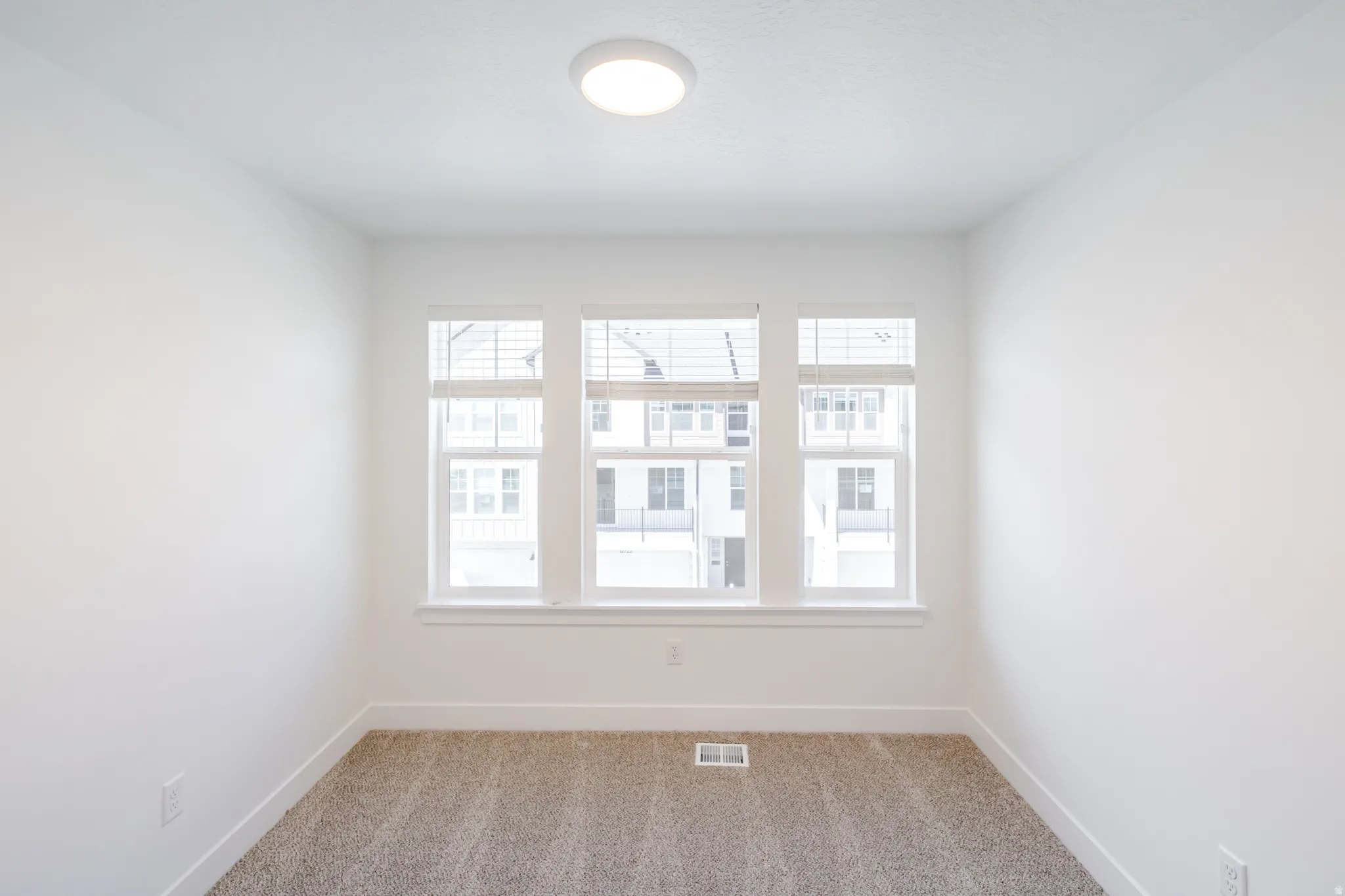 Room featuring three white-framed windows with blinds, light tan carpeting, white baseboards, and a flush-mount ceiling light fixture