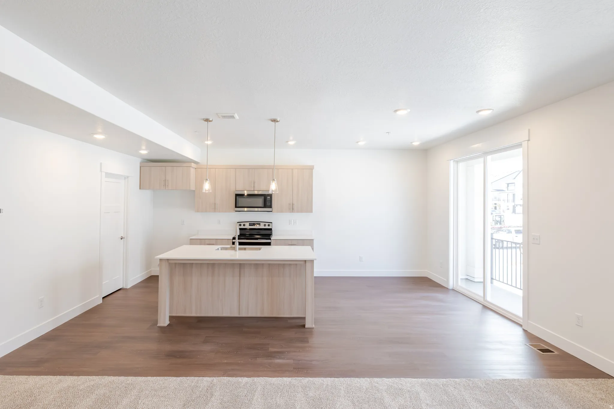 Open-concept living area featuring a kitchen island, wood-finish flooring, recessed lighting, and a sliding glass door leading to a balcony with black railings