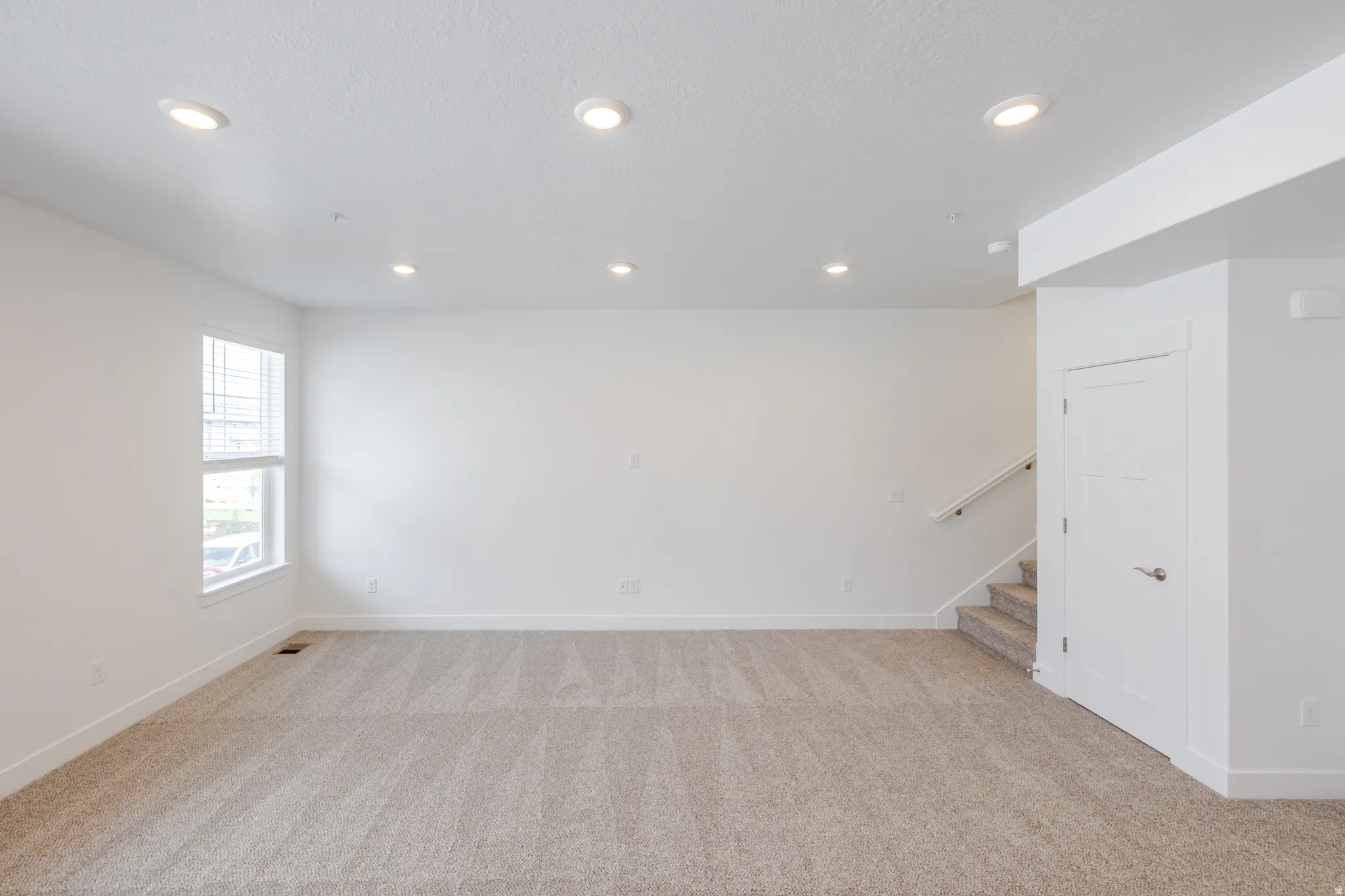 Spacious room featuring neutral carpeting, white walls, recessed lighting, and a prominent window with blinds