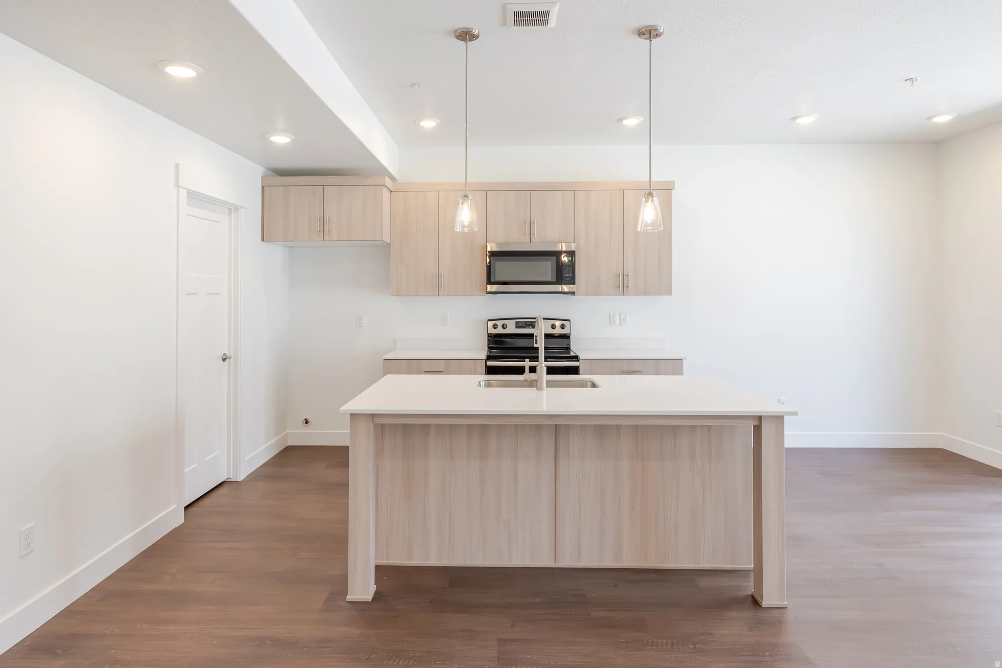 Modern kitchen featuring a central island with an undermount sink and countertop, light wood-finish cabinetry, stainless steel appliances, and wood-finish flooring