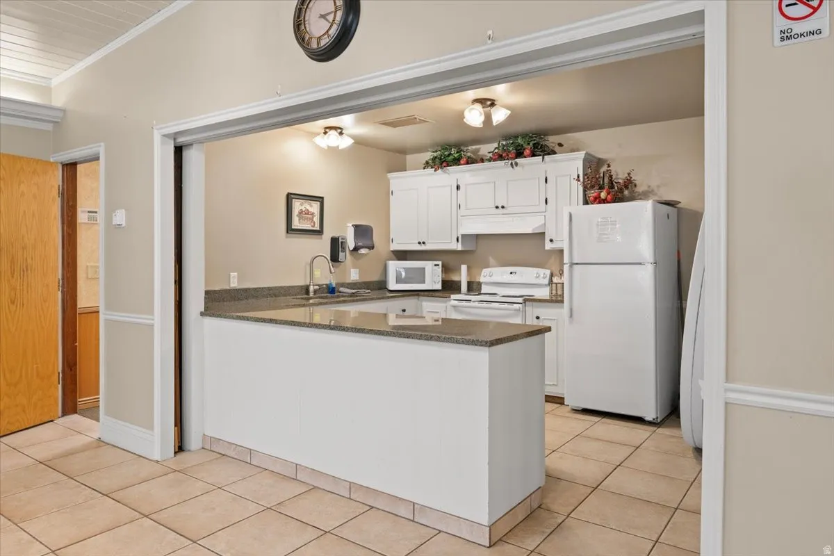 Kitchen with white appliances, a peninsula, white cabinets, dark stone countertops, and ornamental molding