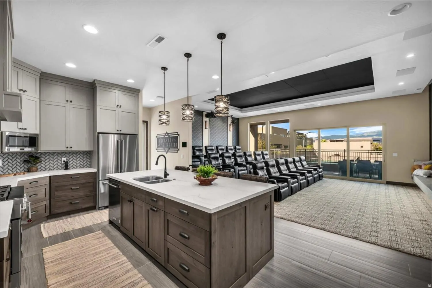 Kitchen with a raised ceiling, stainless steel appliances, hanging light fixtures, a center island with sink, and light stone counters