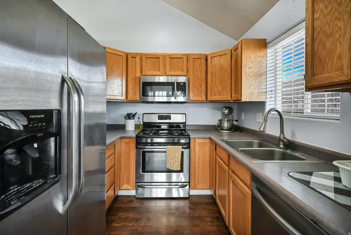 Kitchen featuring stainless steel appliances, vaulted ceiling, dark wood-type flooring, brown cabinets, and dark countertops