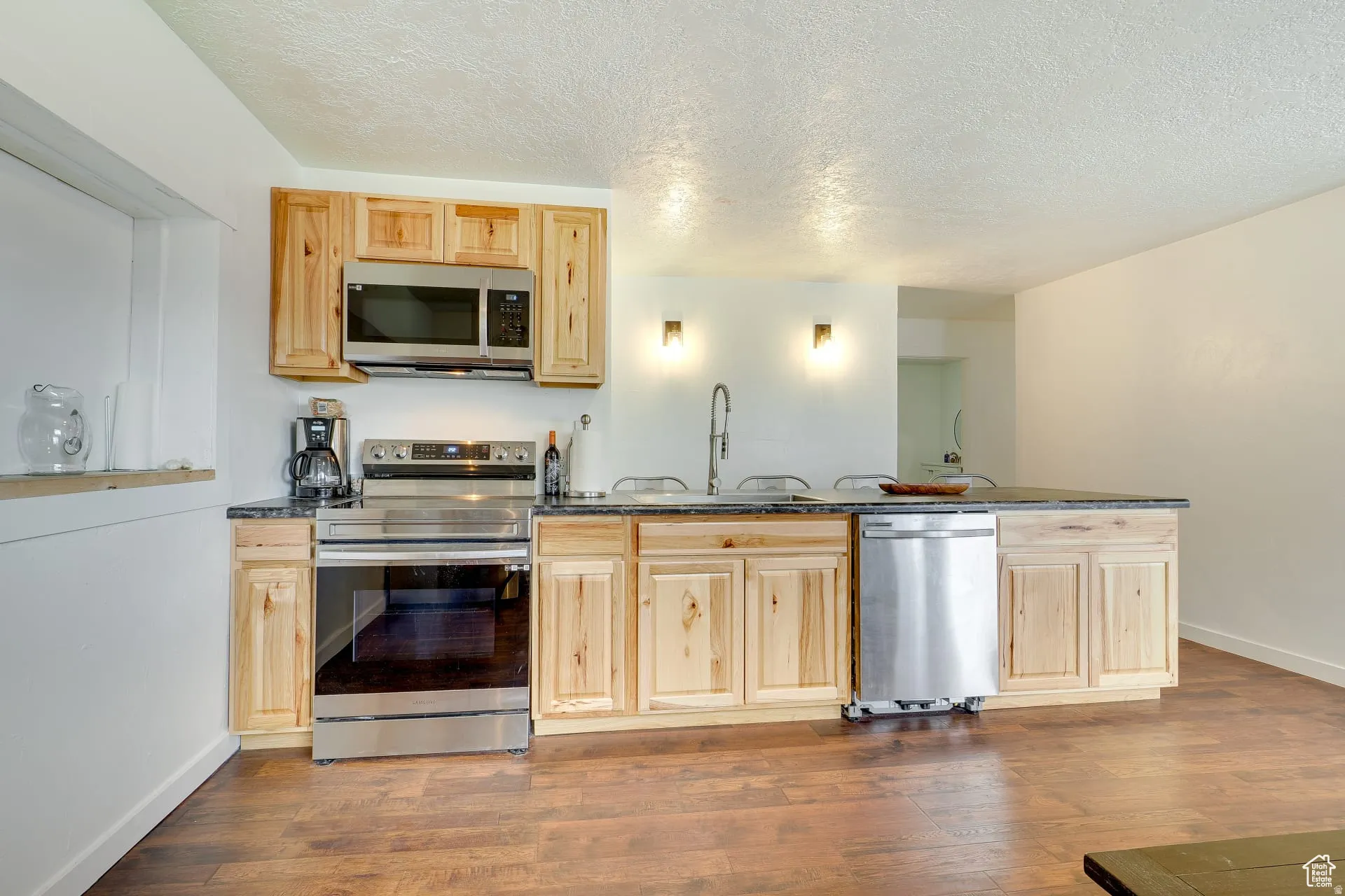 Kitchen featuring stainless steel appliances, light brown cabinets, a textured ceiling, dark stone countertops, and dark wood-style floors