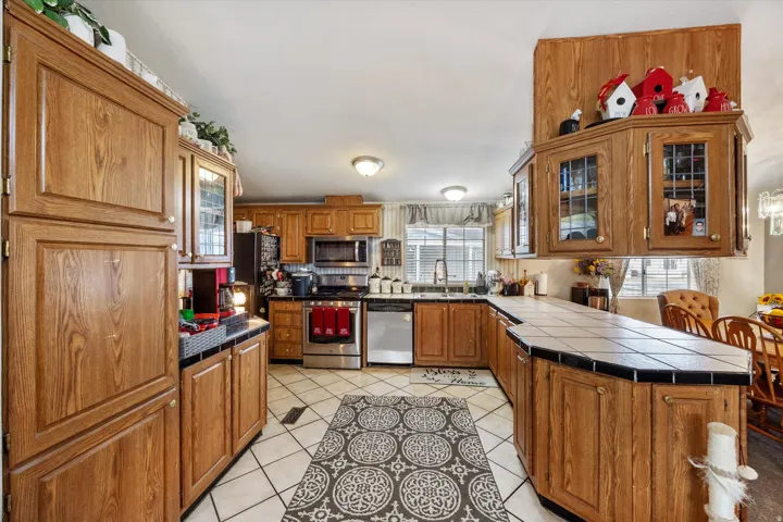 Kitchen with glass insert cabinets, brown cabinets, light tile patterned floors, appliances with stainless steel finishes, and tile counters