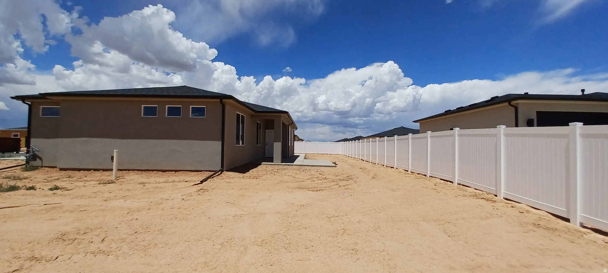 Newly constructed stucco exterior featuring a dark shingled roof, white-framed windows, and a white vinyl privacy fence