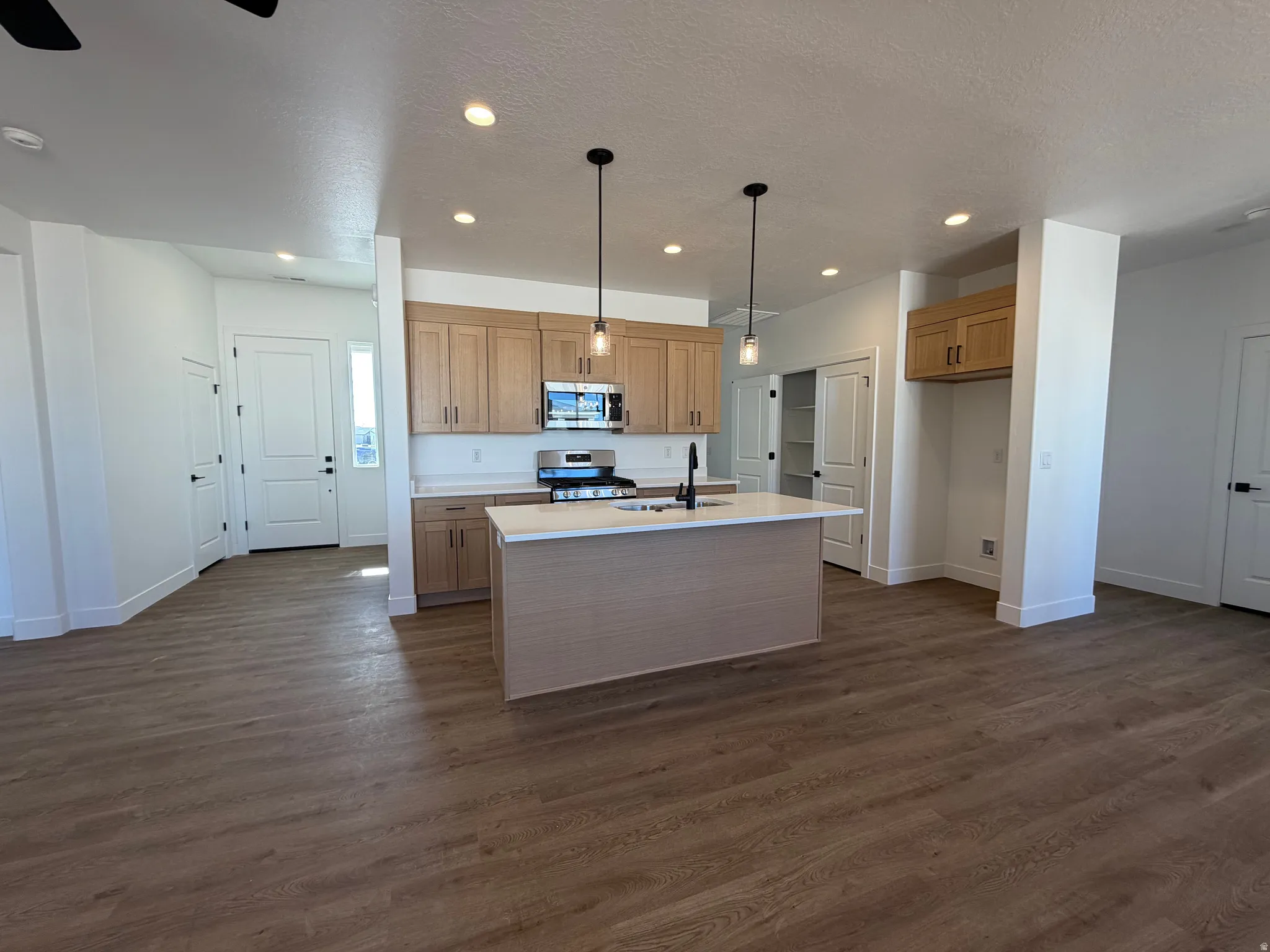Open-concept kitchen featuring light wood cabinetry, stainless steel appliances, a center island with sink and pendant lighting, and light-toned countertops