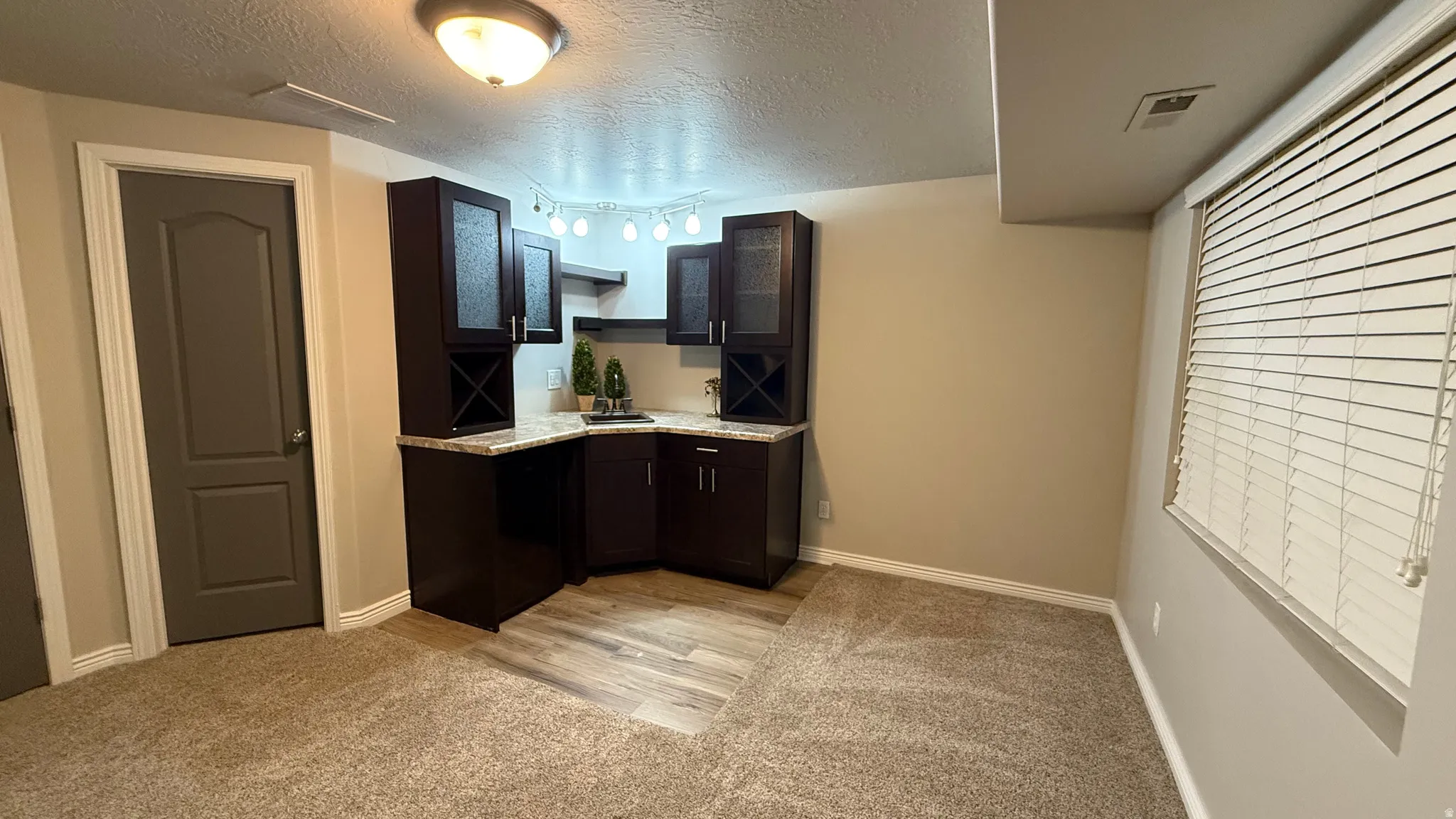 Kitchen with light carpet, a textured ceiling, dark brown cabinetry, light stone countertops, and track lighting