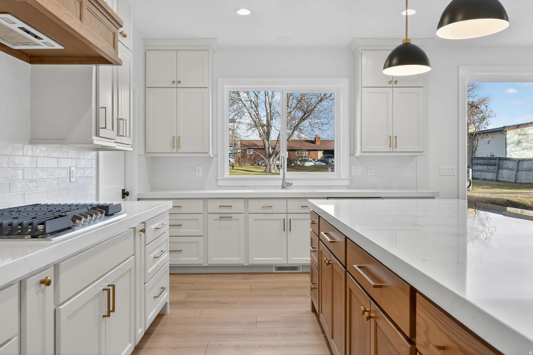 Kitchen featuring off white  rollout cabinetry, premium range hood, light stone countertops, stainless steel gas cooktop, and brown cabinetry, farmhouse sink.