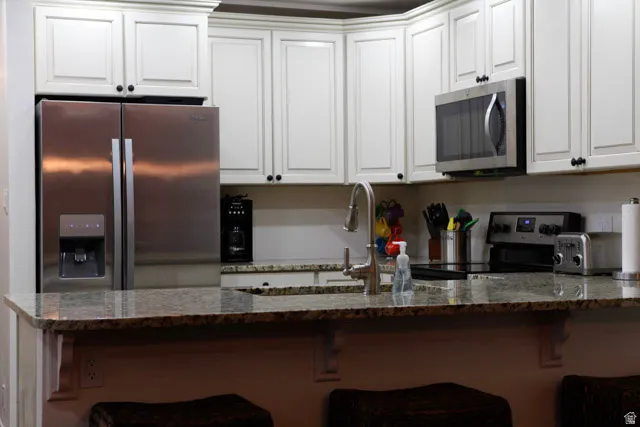 Kitchen featuring dark stone counters, appliances with stainless steel finishes, a kitchen bar, white cabinetry, and a peninsula