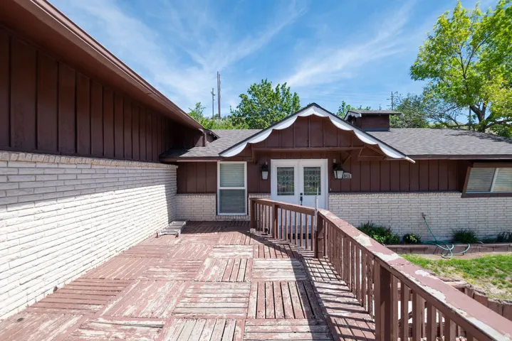Front exterior featuring a wood-finish deck with railing, white brick facade, brown vertical siding, and a gabled entry with double glass-panel doors