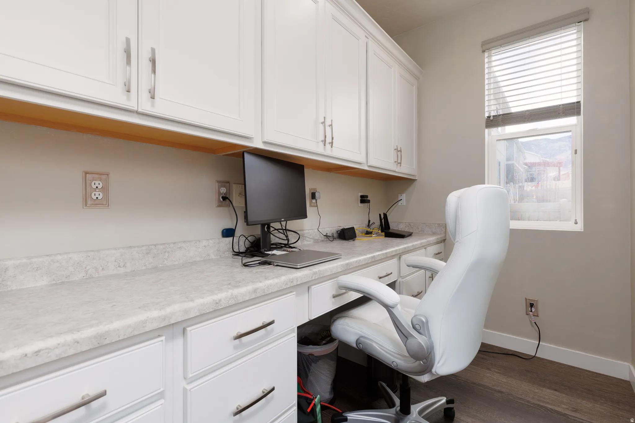 Home office with dark wood-type flooring and baseboards, window with custom blind and expansive work space with cabinetry.