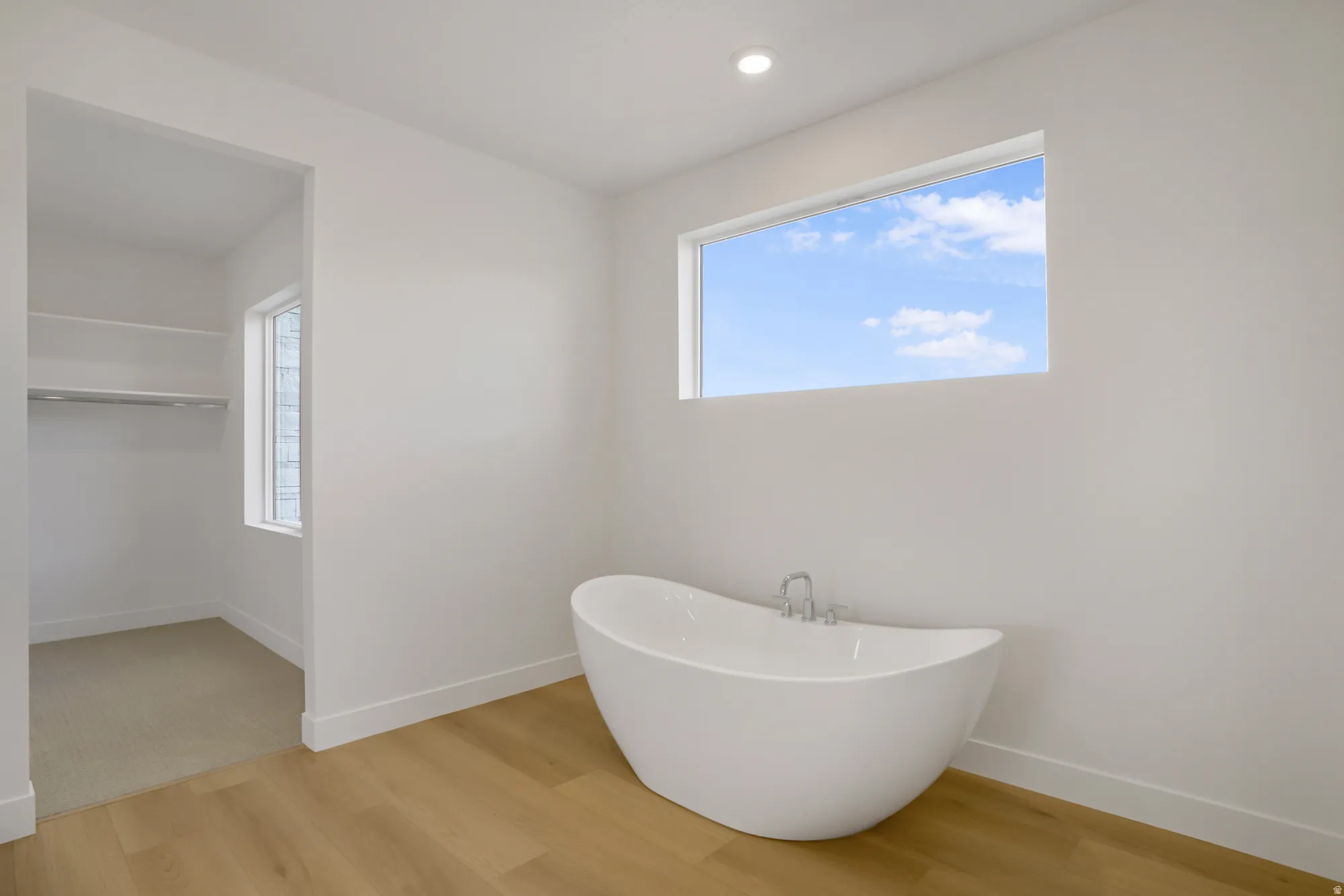 Freestanding soaking tub with polished chrome fixtures, horizontal window, wood-finish flooring, recessed lighting, and an adjacent walk-in closet with built-in shelving