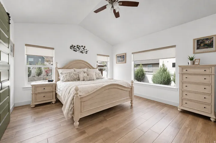 Vaulted ceiling with a ceiling fan, wood-finish flooring, two large windows, white walls, and a sliding barn door