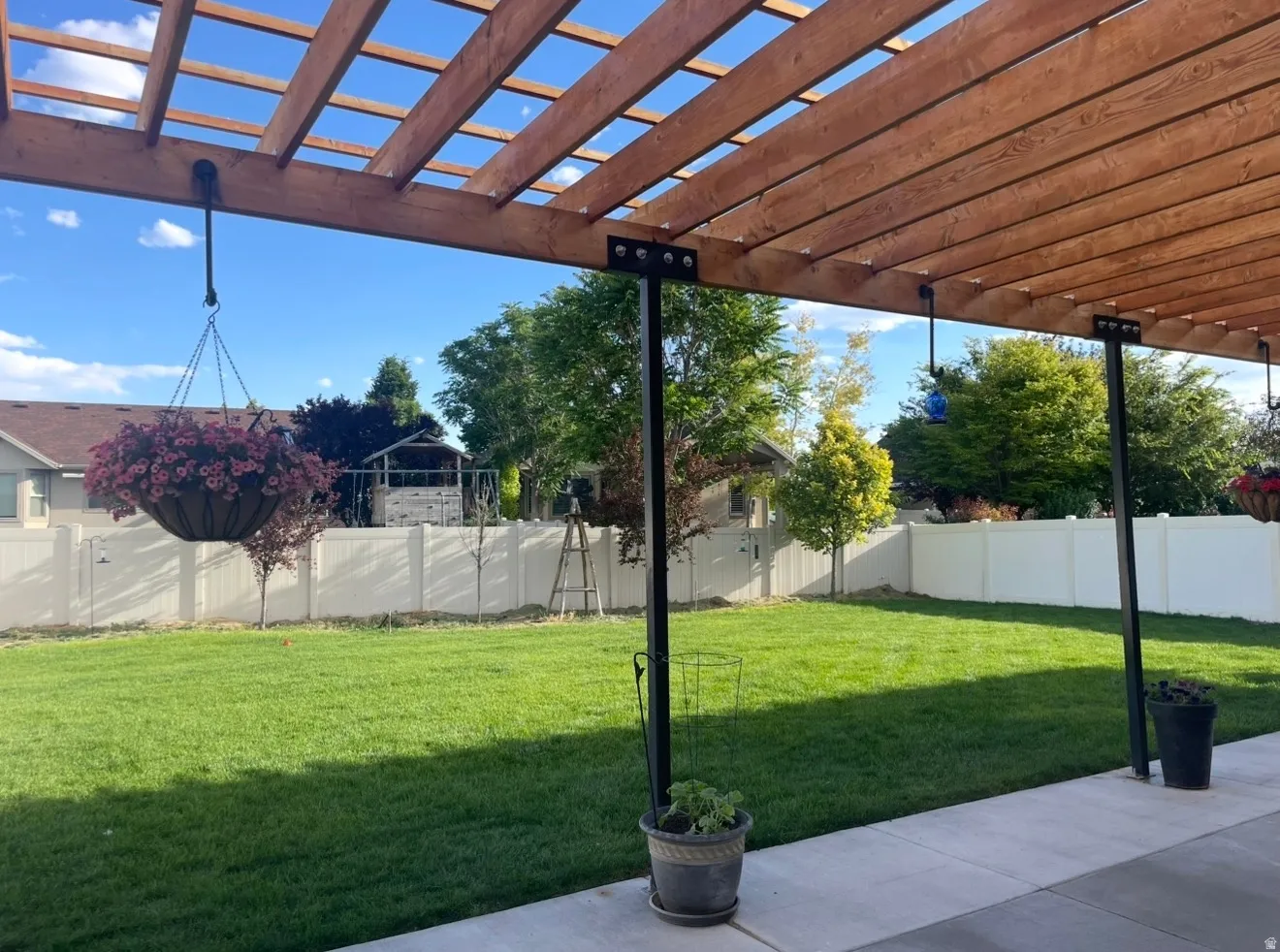 Expansive backyard featuring a wood pergola with metal supports, concrete patio, lush green lawn, and white privacy fencing