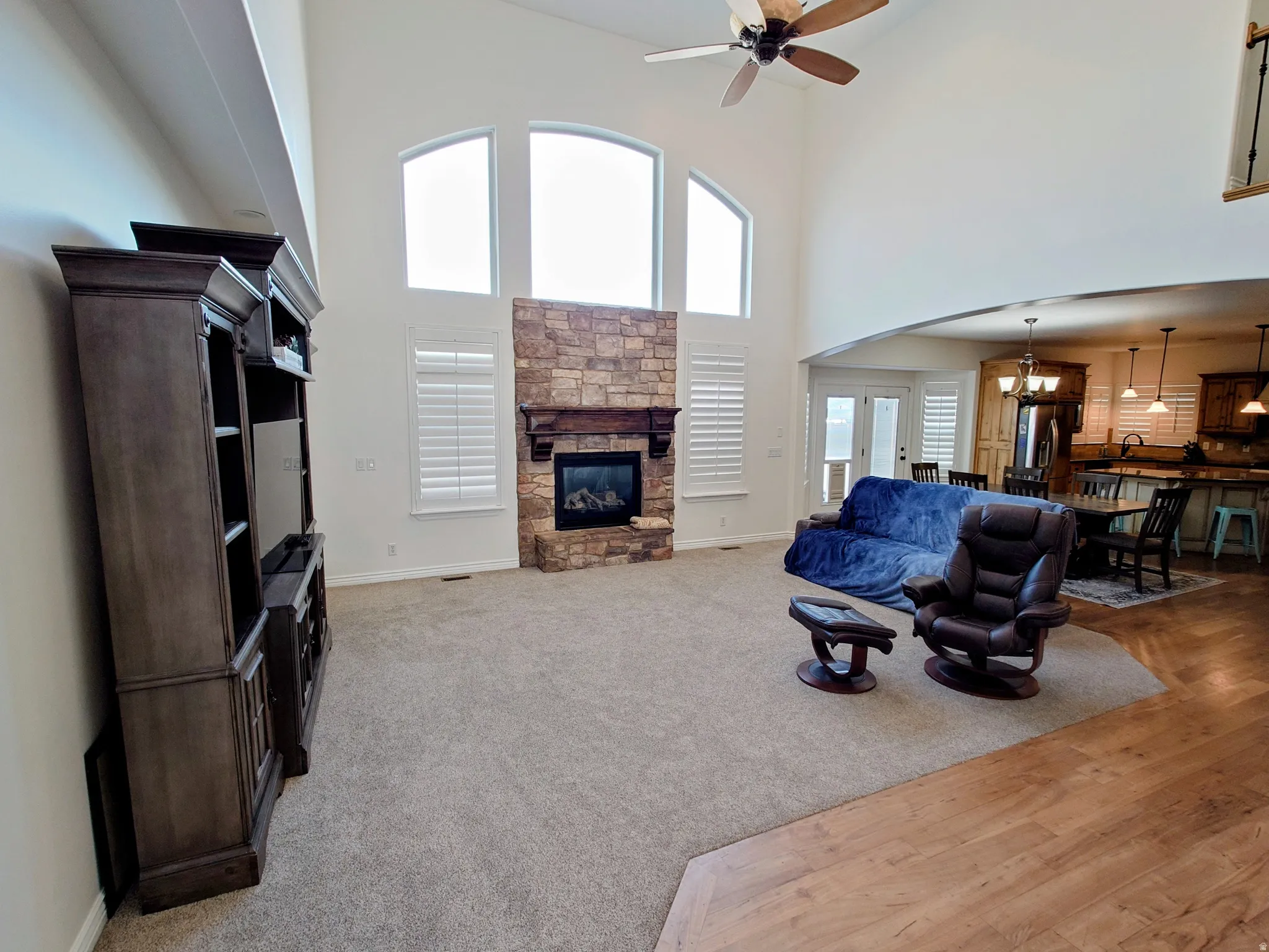 Living room featuring a high ceiling, a stone fireplace, ceiling fan, a chandelier, and light colored carpet