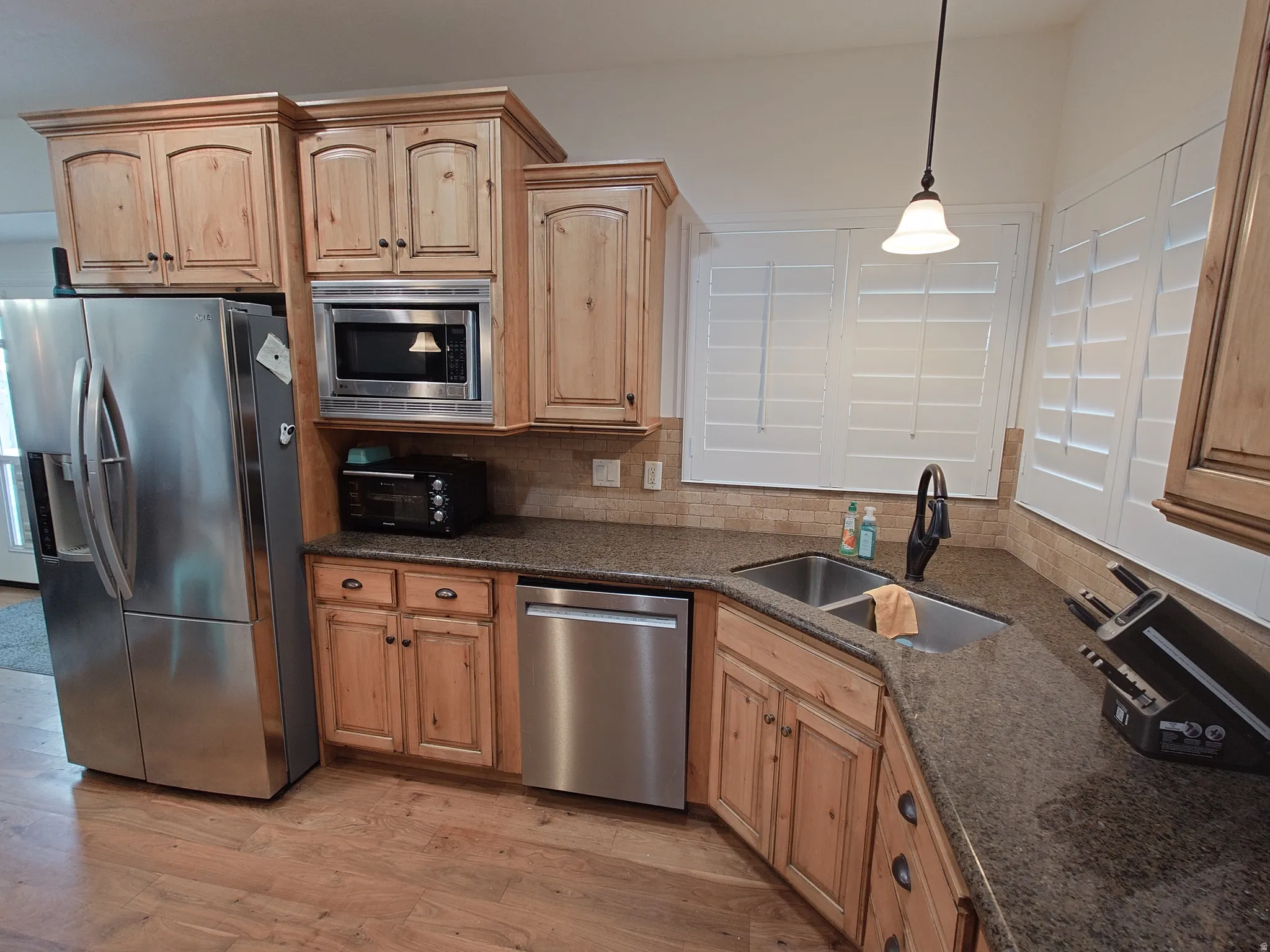 Kitchen with stainless steel appliances, backsplash, light wood-type flooring, dark stone countertops, and decorative light fixtures