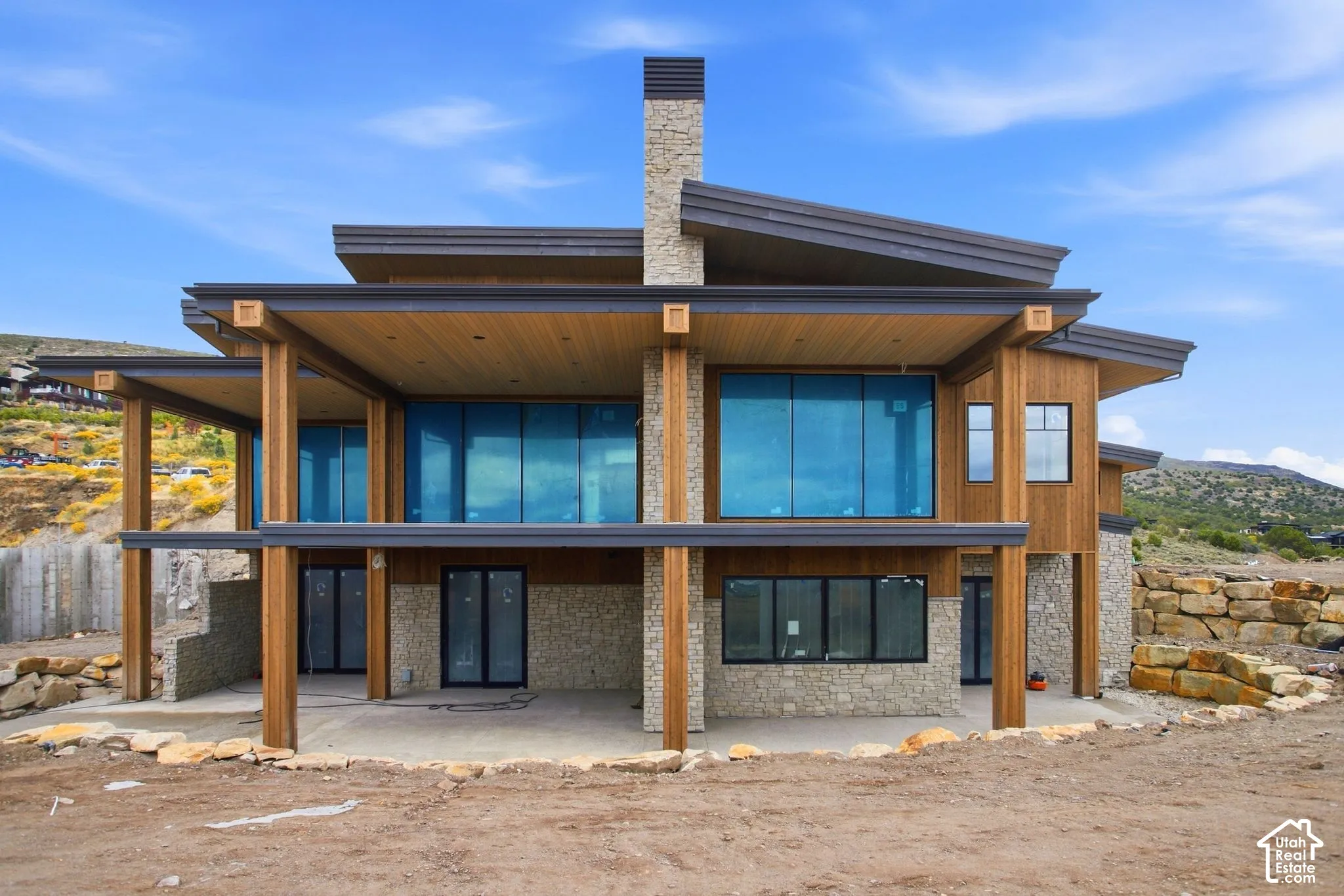 Modern architectural design featuring extensive glazing, stone and wood panel exterior, a prominent stone chimney, and a multi-level covered patio