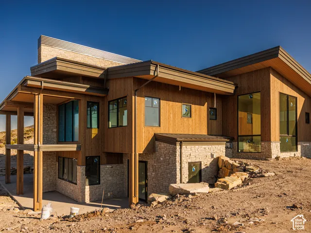 Modern architectural exterior featuring vertical wood-finish siding, stacked stone masonry, expansive floor-to-ceiling windows, deep roof overhangs, and a covered lower-level patio