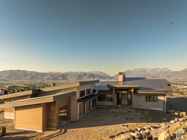 Modern residence featuring a multi-tonal stone facade, horizontal wood siding, standing-seam metal roofing, and a prominent stone chimney, set against a backdrop of mountain peaks