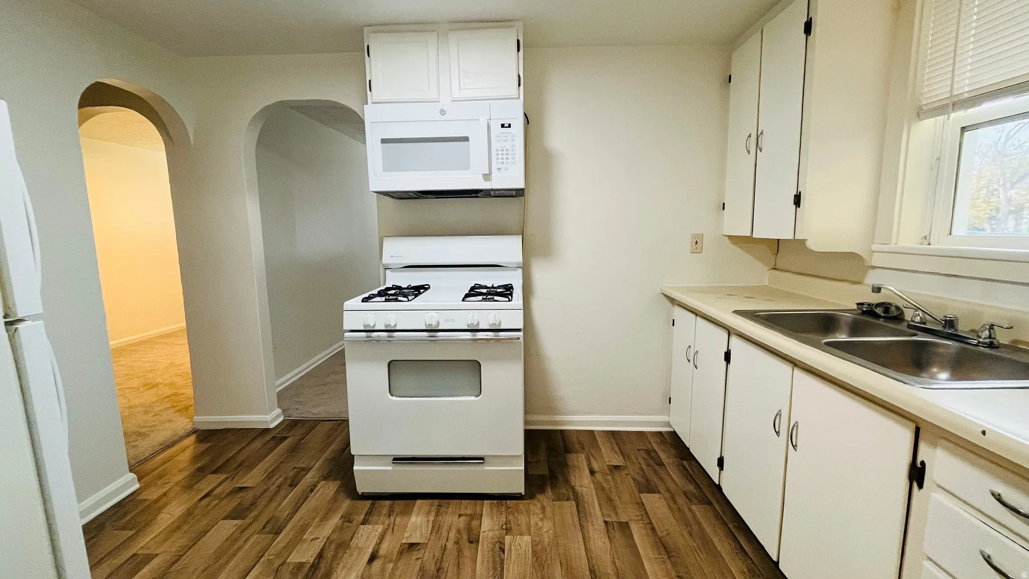 Kitchen featuring arched walkways, white appliances, white cabinetry, light countertops, and dark wood-type flooring