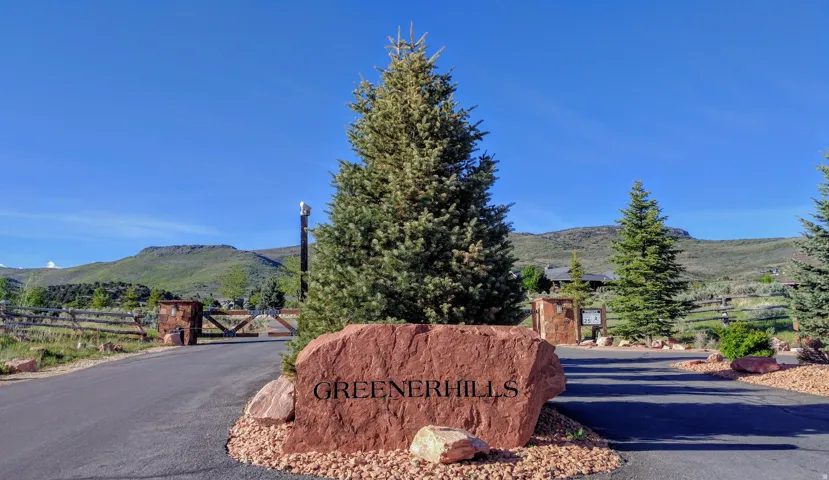 Paved entrance featuring a "Greenerhills" stone sign, mature evergreen trees, and a distant mountainous backdrop