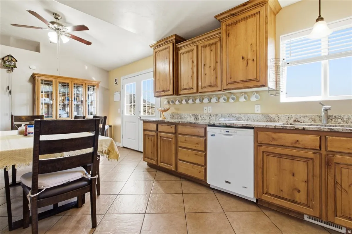 Kitchen featuring white dishwasher, brown cabinetry, light stone counters, decorative light fixtures, and lofted ceiling