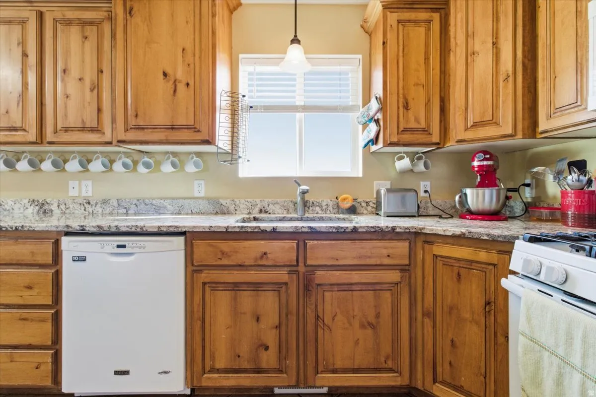 Kitchen with brown cabinetry, white appliances, hanging light fixtures, and light stone countertops