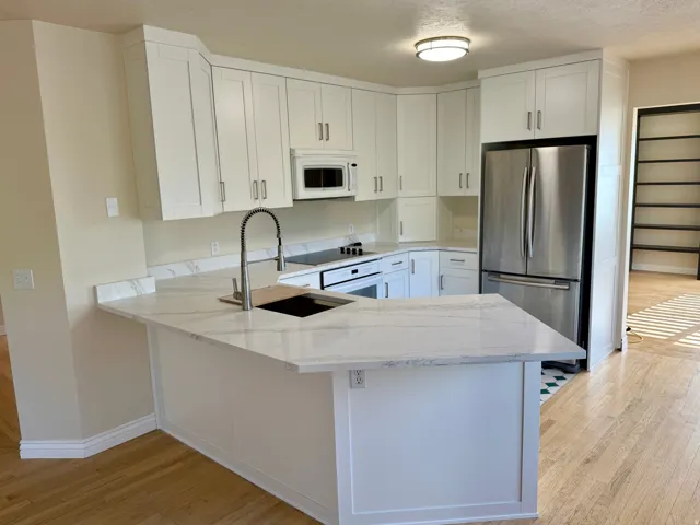 Modern kitchen featuring white shaker cabinetry, stainless steel appliances, a corner sink with a gooseneck faucet, light-toned wood-finish flooring, and a stone-finish countertop island with seating space