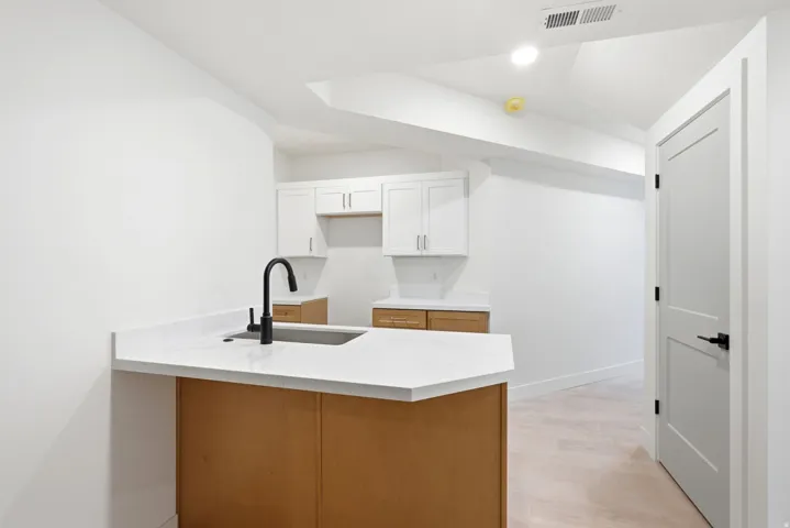 Kitchenette featuring a stainless steel sink, black gooseneck faucet, light wood-finish cabinetry, white upper cabinets, and white countertops