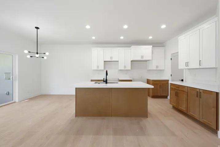 Kitchen featuring white upper cabinetry, wood-finish lower cabinetry, a center island with sink, black faucet, and light wood-finish flooring