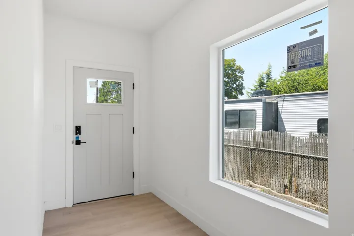 White paneled entry door with glass insert, light wood-finish flooring, and a large window with white trim