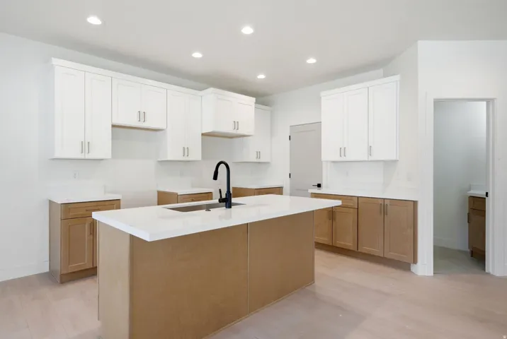 Kitchen featuring an island with an integrated sink and matte black faucet, light wood-finish flooring, white upper cabinetry, and natural wood-finish lower cabinetry, complemented by recessed lighting