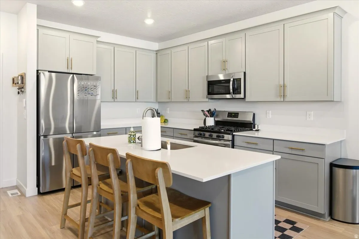 Kitchen featuring stainless steel appliances, gray cabinetry, a breakfast bar, light wood-type flooring, and a center island with sink