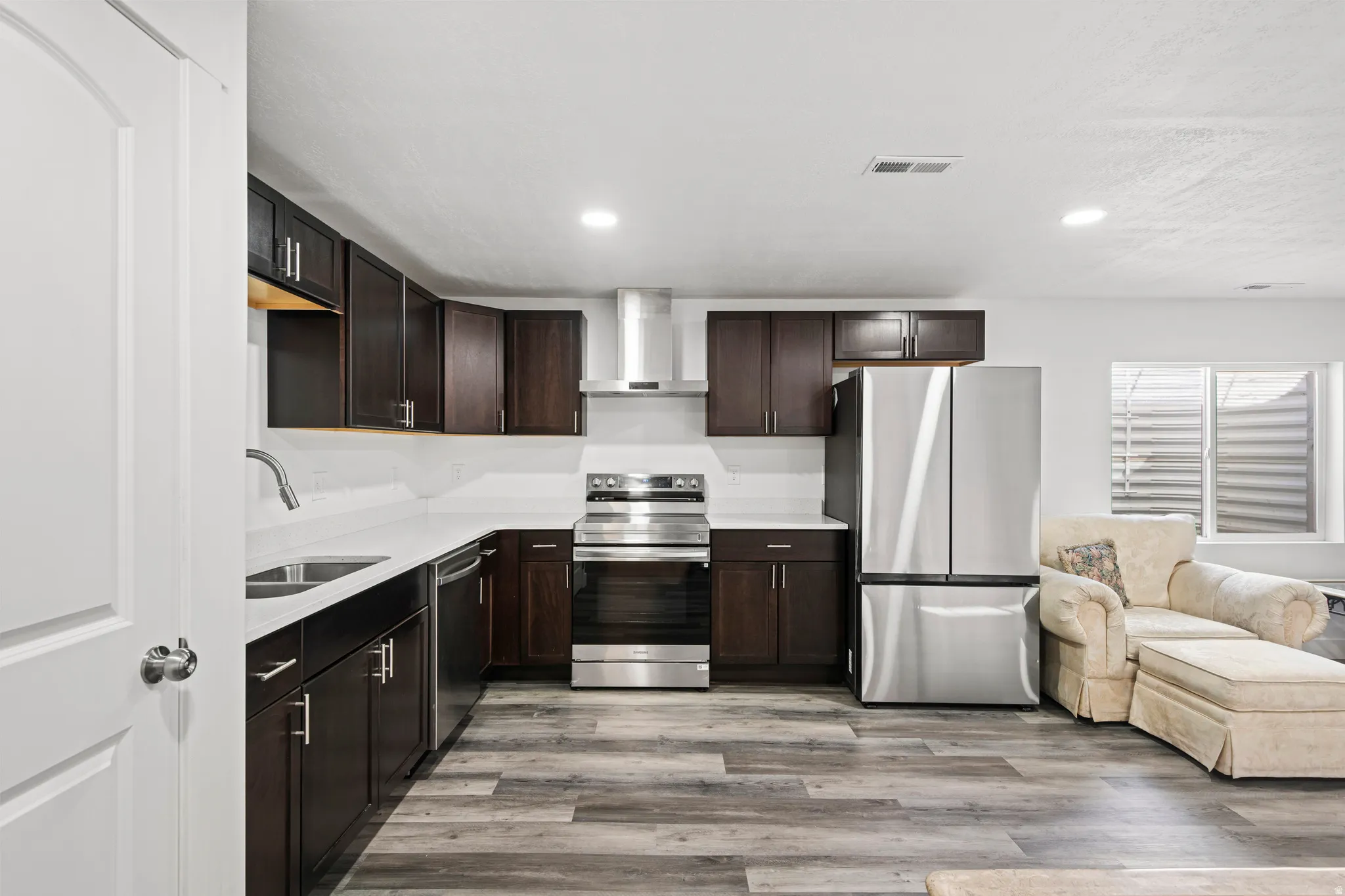 Kitchen with appliances with stainless steel finishes, dark brown cabinetry, recessed lighting, light wood-type flooring, and wall chimney range hood