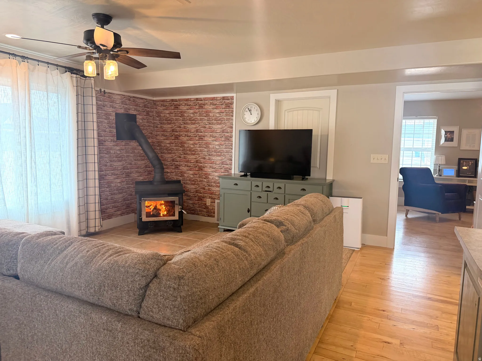 Living room with a wood stove, brick wall, light wood-type flooring, and ceiling fan