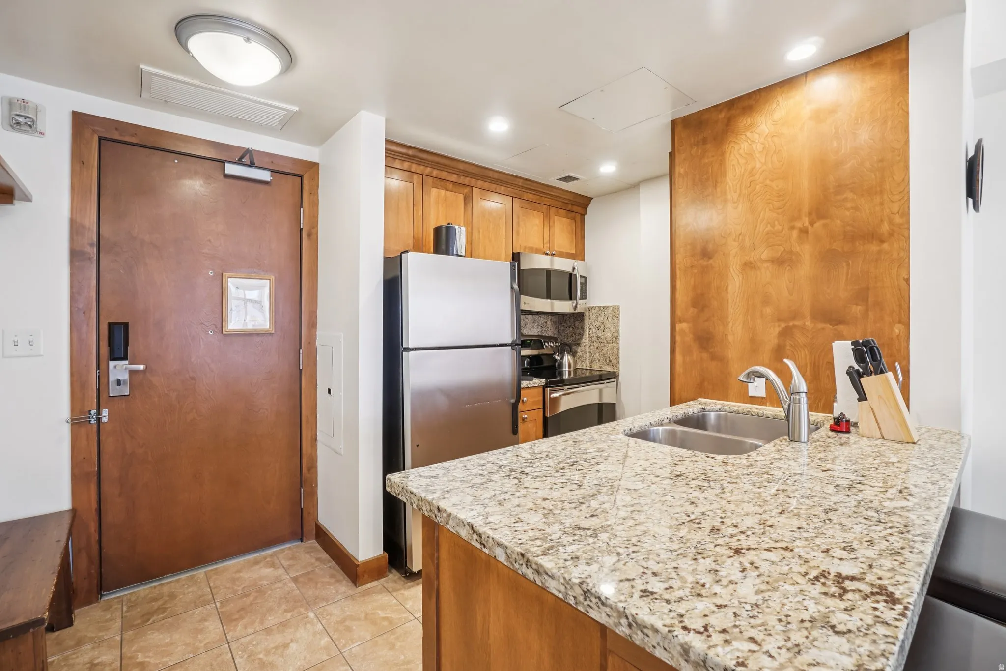 Kitchen featuring brown cabinetry, appliances with stainless steel finishes, a peninsula, light stone counters, and light tile patterned flooring