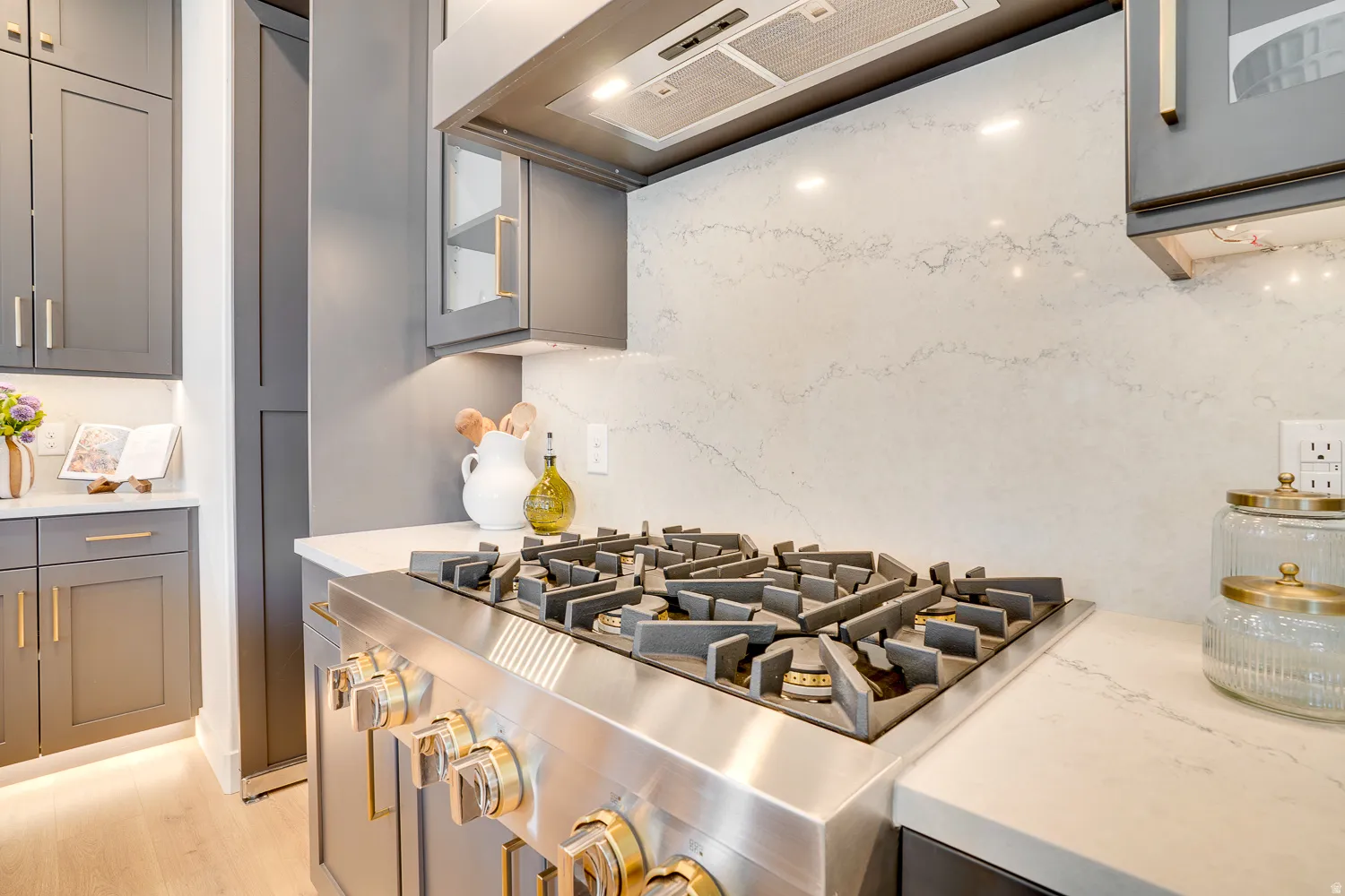 Kitchen view of ventilation hood, gray cabinetry, light stone counters, and glass insert cabinets