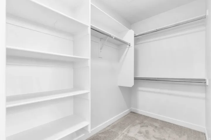 Spacious walk-in closet featuring white built-in shelving, chrome hanging rods, and light gray carpeting