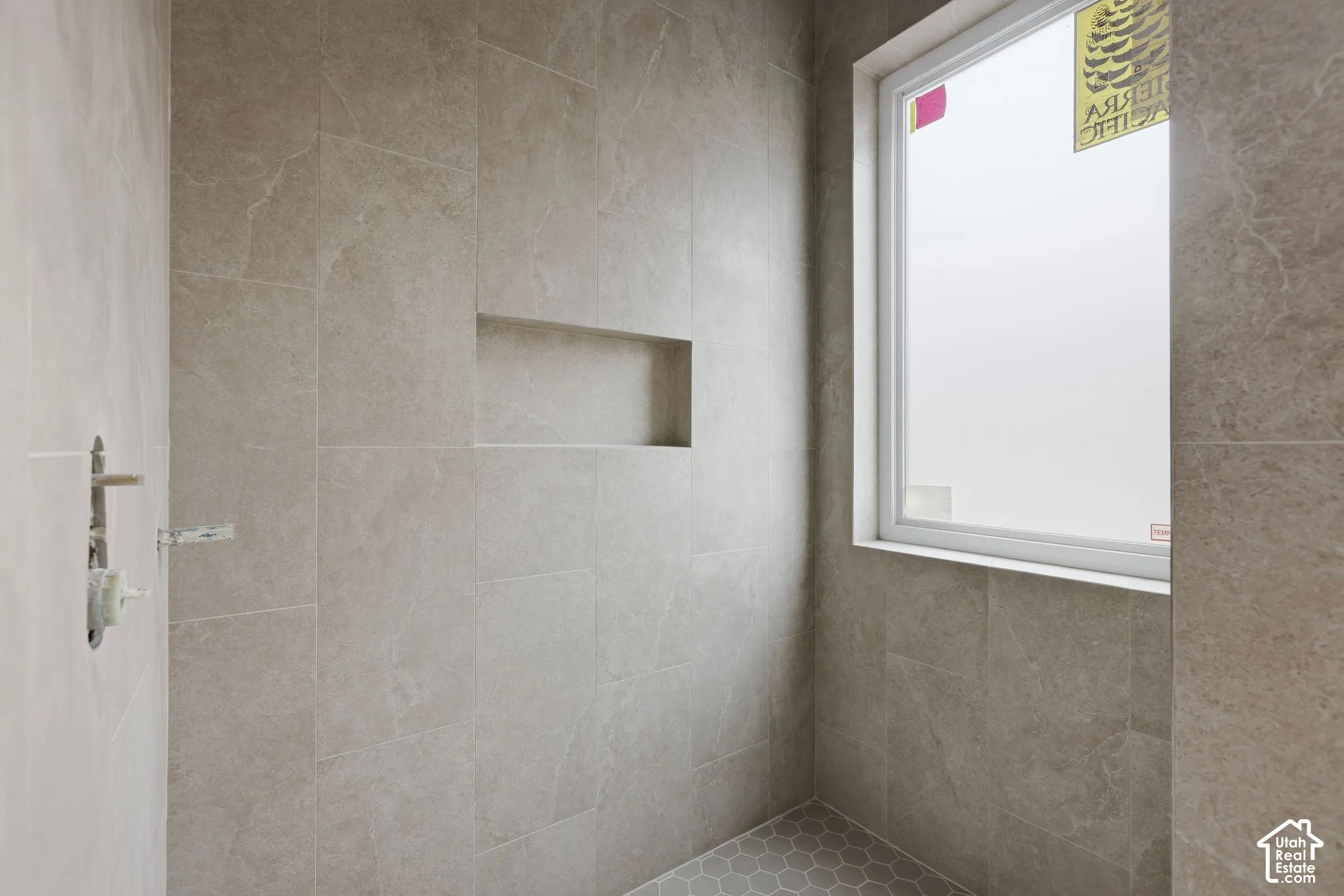 Shower enclosure featuring large-format neutral-toned wall tile, a recessed storage niche, a single-pane window, and hexagonal floor tiling