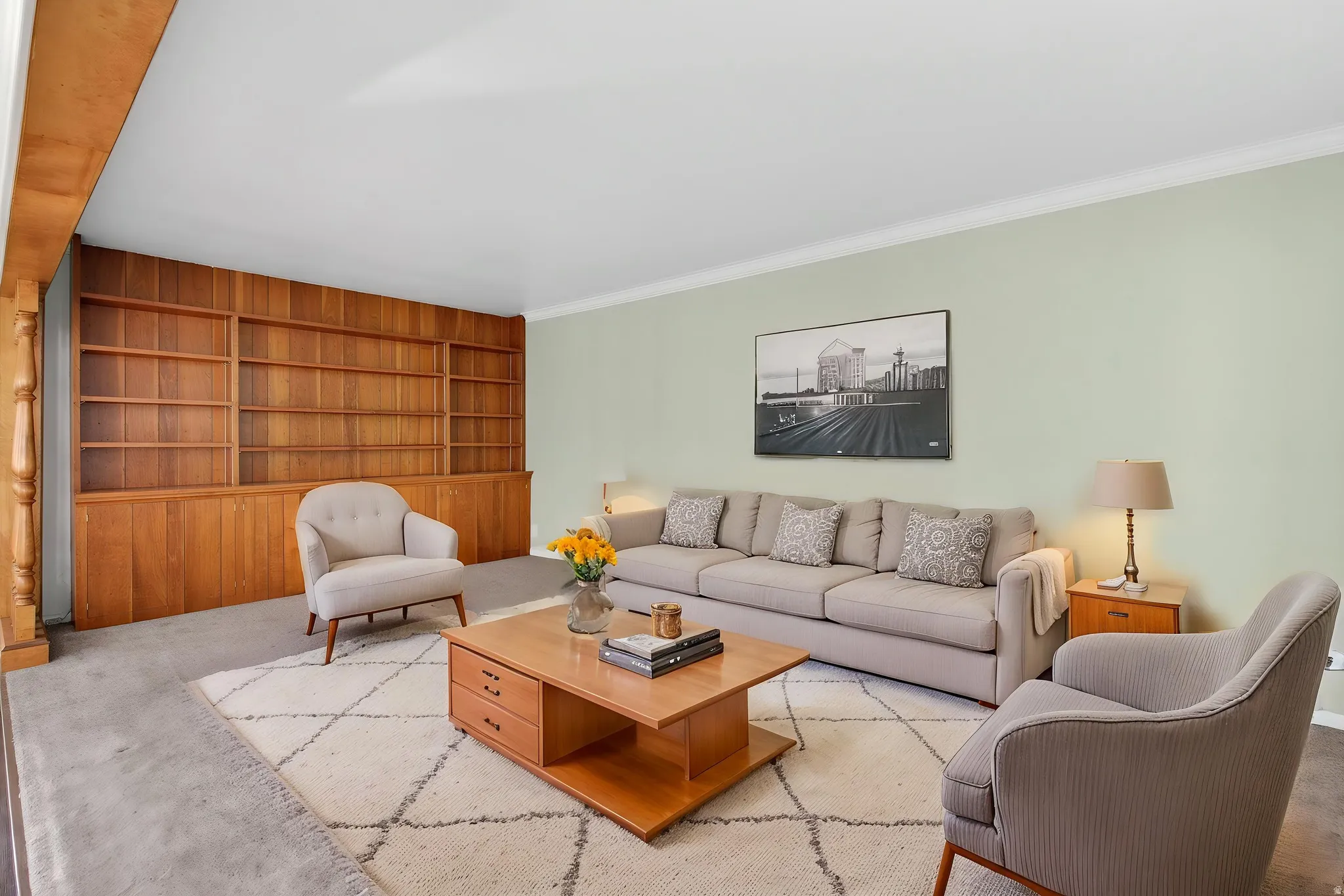 Virtual staging suggestion. Large living room with integrated wood-paneled shelving and built-in cabinetry, crown molding, light-toned wall color, and carpeted flooring.