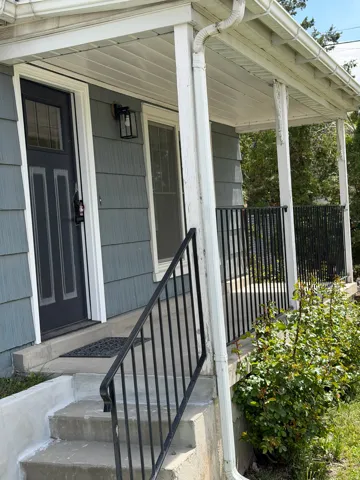 Covered front porch with a dark entry door, white trim, exterior wall sconce, concrete steps, and black metal railing