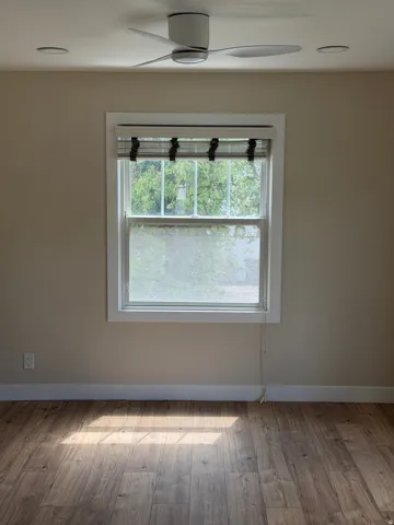 Room featuring wood-finish flooring, a window with a fabric Roman shade, and a ceiling fan with integrated lighting