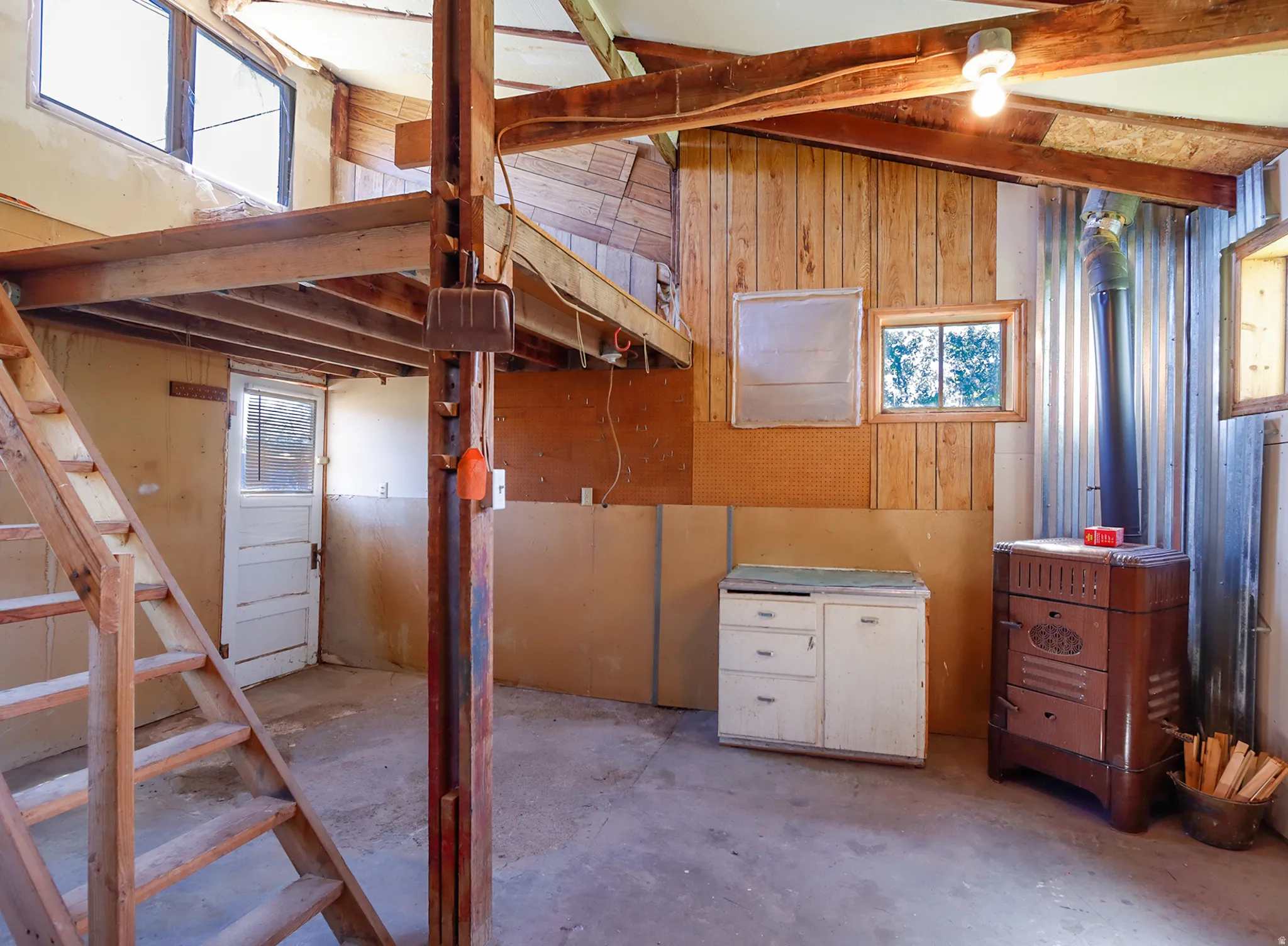 Interior space featuring exposed wood beams, a loft area with wood stairs, a built-in wood-burning stove with a metal flue, wood paneling, and multiple windows