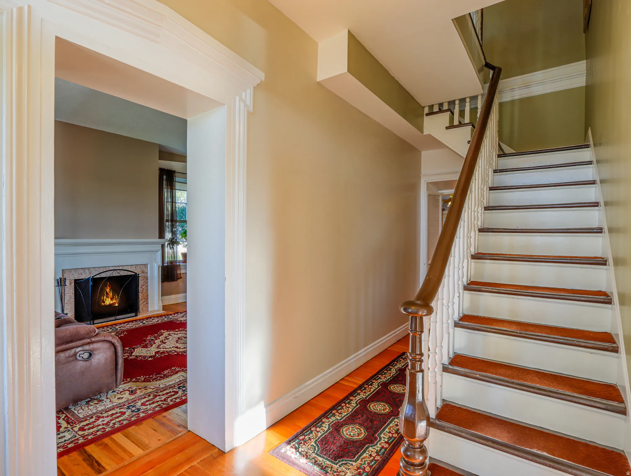 Foyer with wood-finish flooring, a grand staircase with wood newel post and handrail, and an arched doorway framing a living space with a fireplace featuring a stone surround